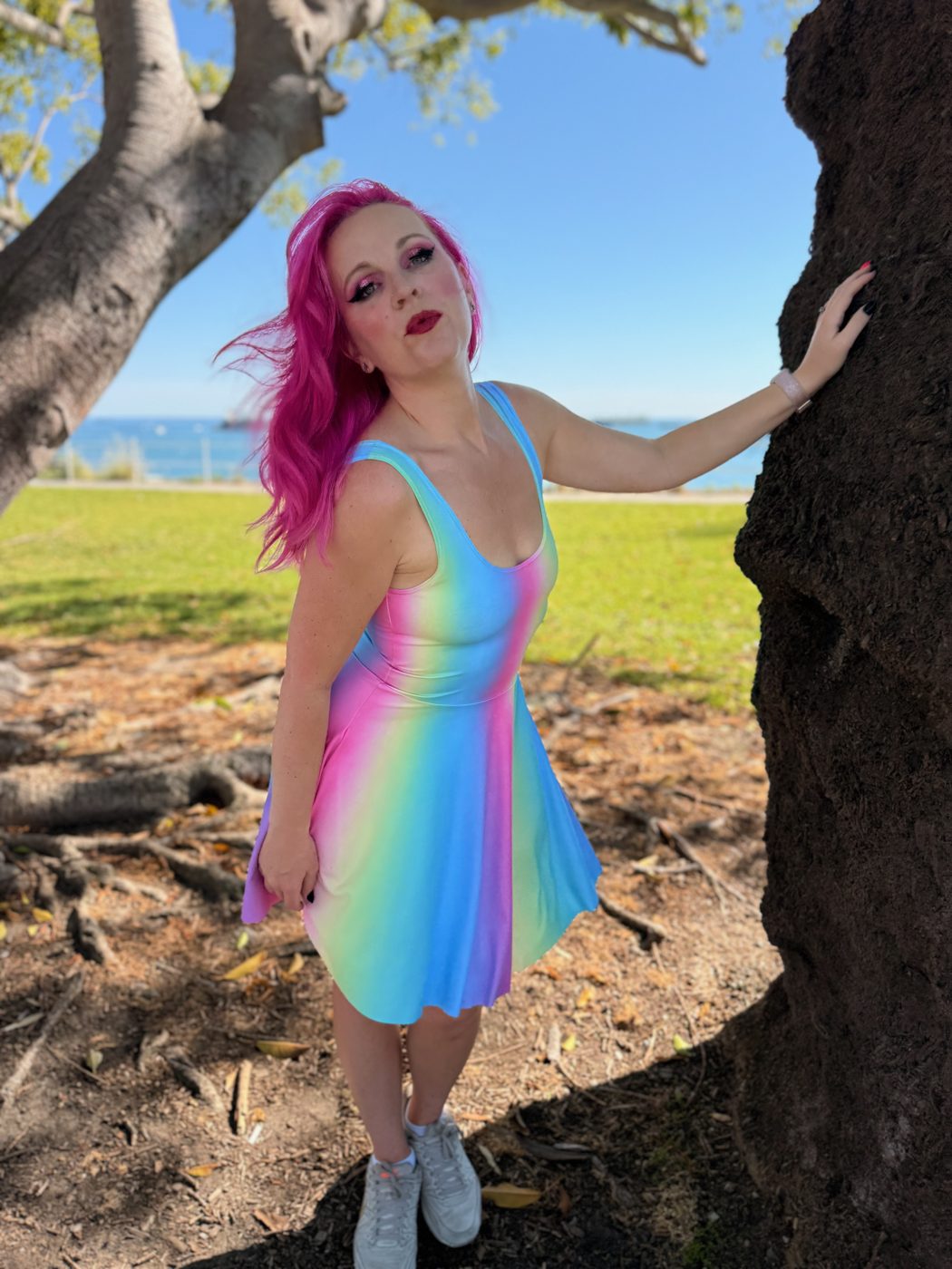 Jessica in a rainbow dress, pink hair, standing by a tree